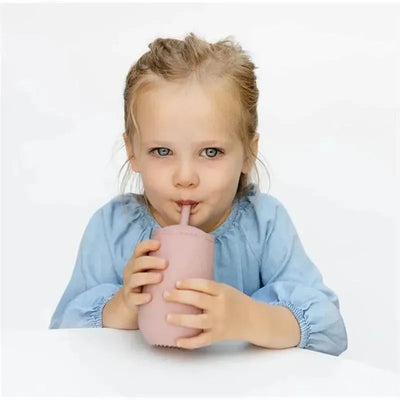 A young girl with blue eyes holds a blush-colored Ezpz Happy Cup with a straw, sipping from it.