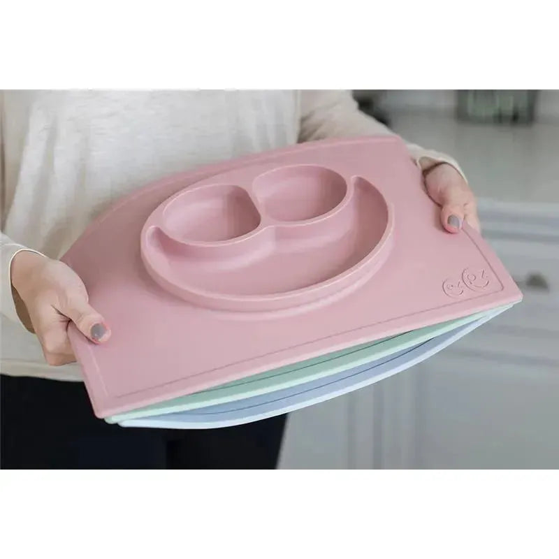 A person holds the Ezpz Happy Mat in blush, featuring a smiley face design and two food compartments, with pastel mats below.