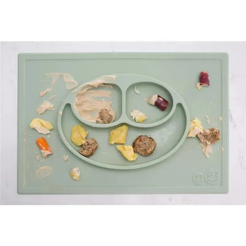 Ezpz Happy Mat in sage color with scattered food items, including meatballs and vegetables on a messy surface.