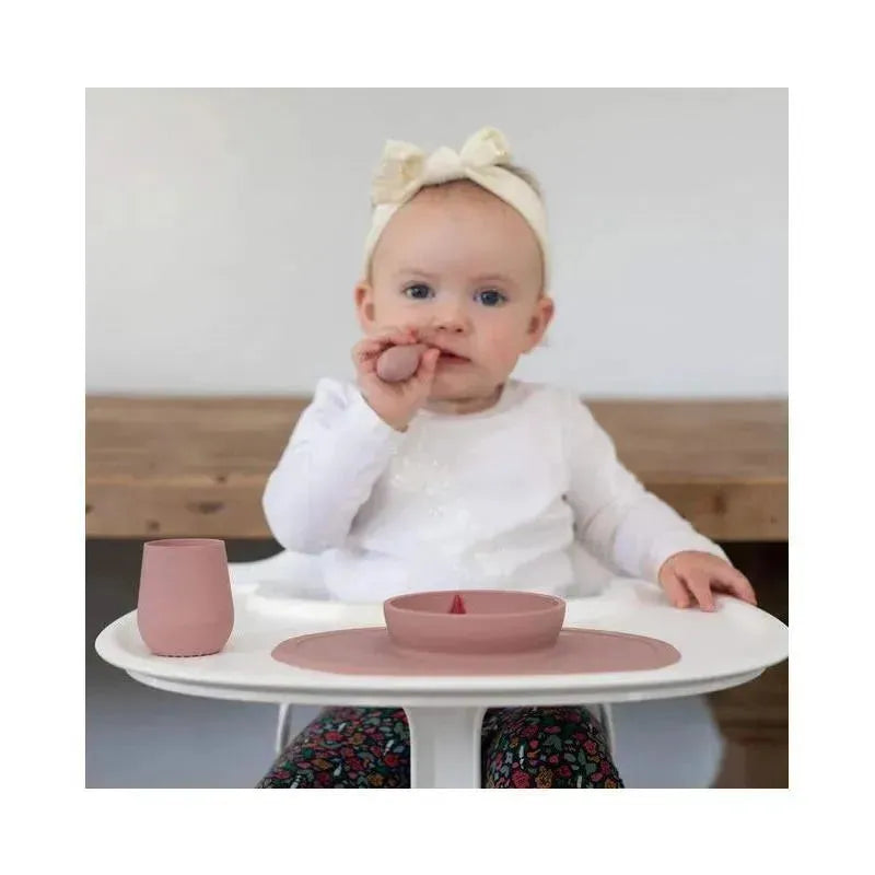 A baby sits at a high chair with a blush-colored Ezpz Tiny Spoon, a matching cup, and a divided plate, holding food.