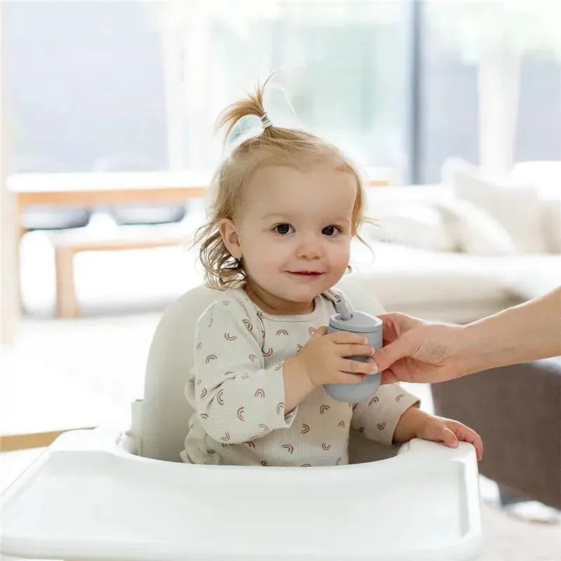 Toddler in a high chair holds a gray Ezpz Mini Cup & Straw Training System, smiling at an adult handing it to them.