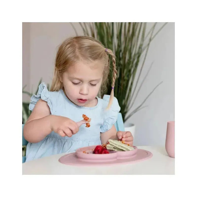 Girl in a light blue dress uses a fork to eat from a blush-colored mini feeding set, featuring strawberries and a sandwich.
