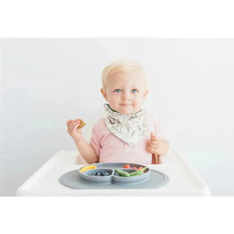 A child wearing a floral bib sits at a high chair, holding a snack, with a plate of fruits on the tray.