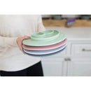 Four colorful silicone plates are stacked, held by a person, showcasing the Ezpz Mini Mat in blush, mint, pink, and gray.