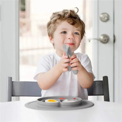 Child holding pewter mini utensils, smiling while seated at a table with a divided plate of food.