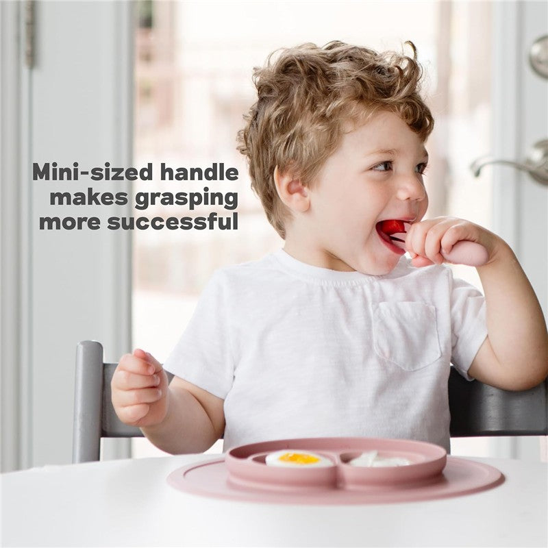 Mini utensils in sage color are used by a child with curly hair, eating from a divided plate that holds food.