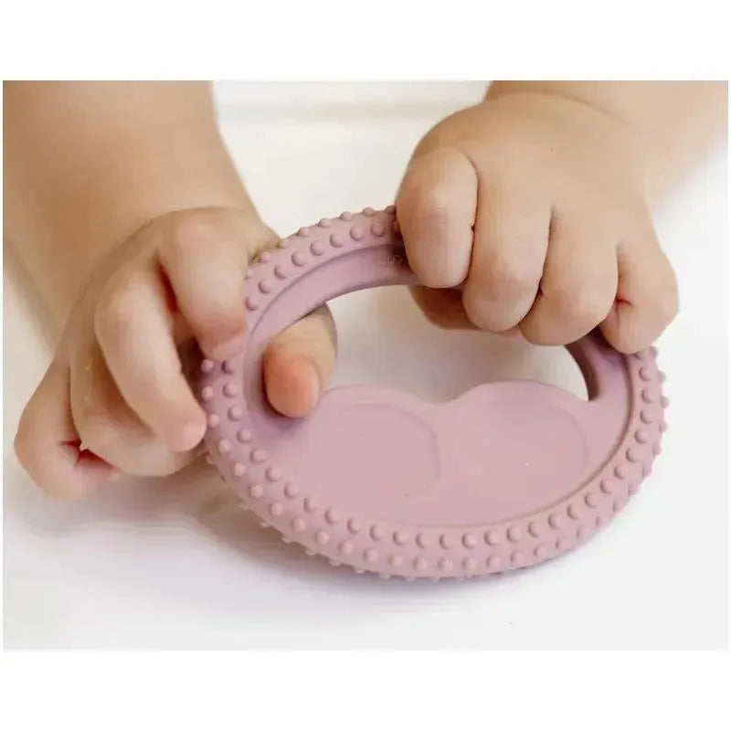 Baby hands grasp a blush-colored, textured silicone oral development tool designed for teething.