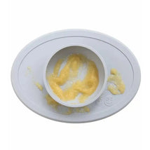 Ezpz Tiny Bowl in pewter color, featuring a curved edge, with a portion of yellow food inside and some spilled around.