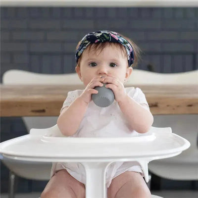 A baby sits in a high chair, holding an Ezpz Tiny Cup in blush. The child has a floral headband and looks curiously at the camera.
