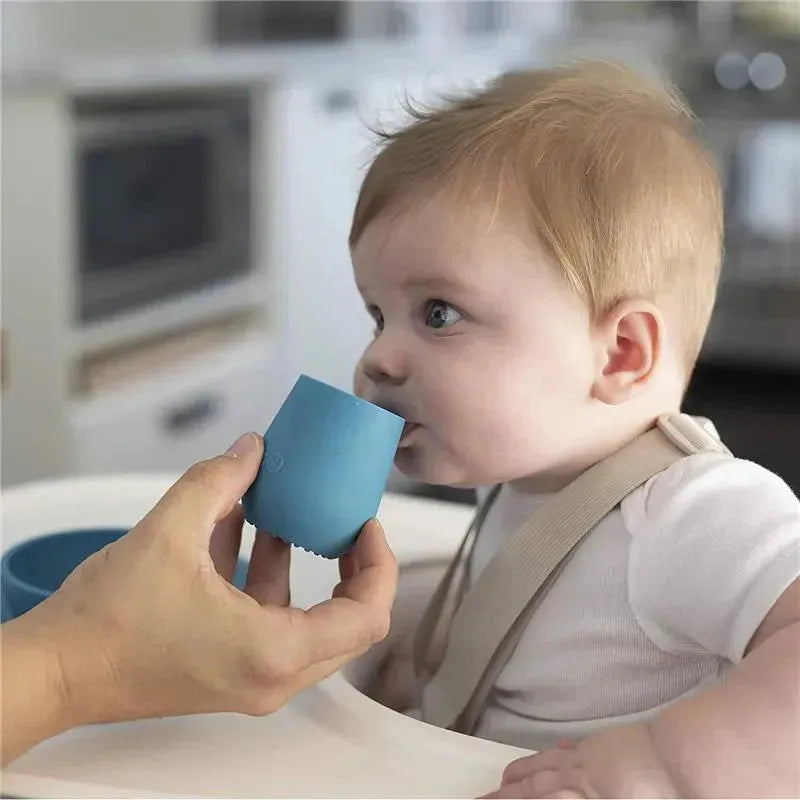 A baby in a high chair is sipping from an Ezpz Tiny Cup in blush, held by an adult hand.