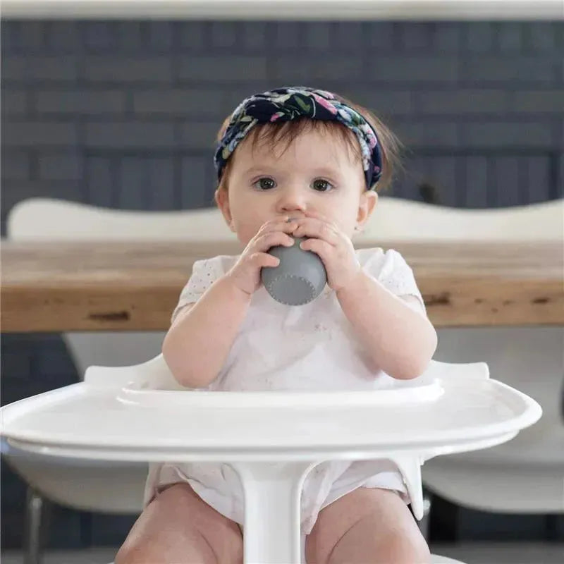 Baby sitting in a high chair, holding a gray Ezpz Tiny Cup, wearing a floral headband and a white outfit.