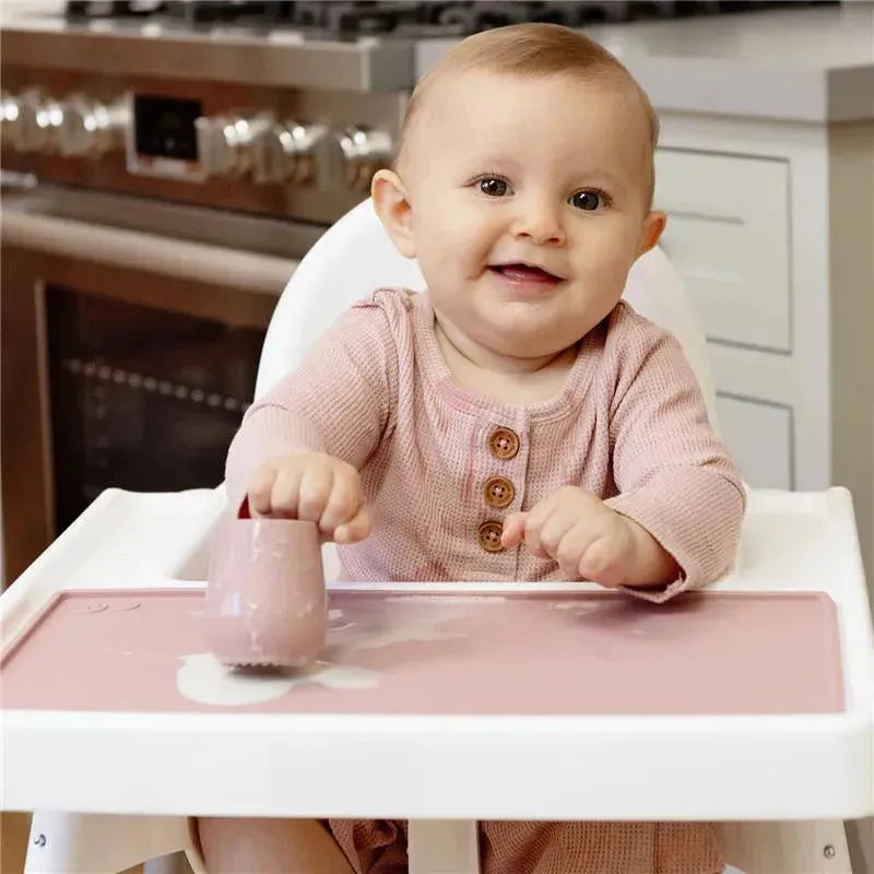 A smiling baby in a high chair plays with a light pink Ezpz Tiny Placemat, featuring a mess-free design.