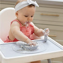 Baby in a high chair plays with a gray Ezpz Tiny Placemat, covered in food, while using a spoon and holding a bowl.
