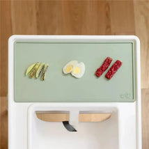 Tiny placemat in sage color features sliced kiwi, halved boiled egg, and two pieces of red food, set on a white surface.