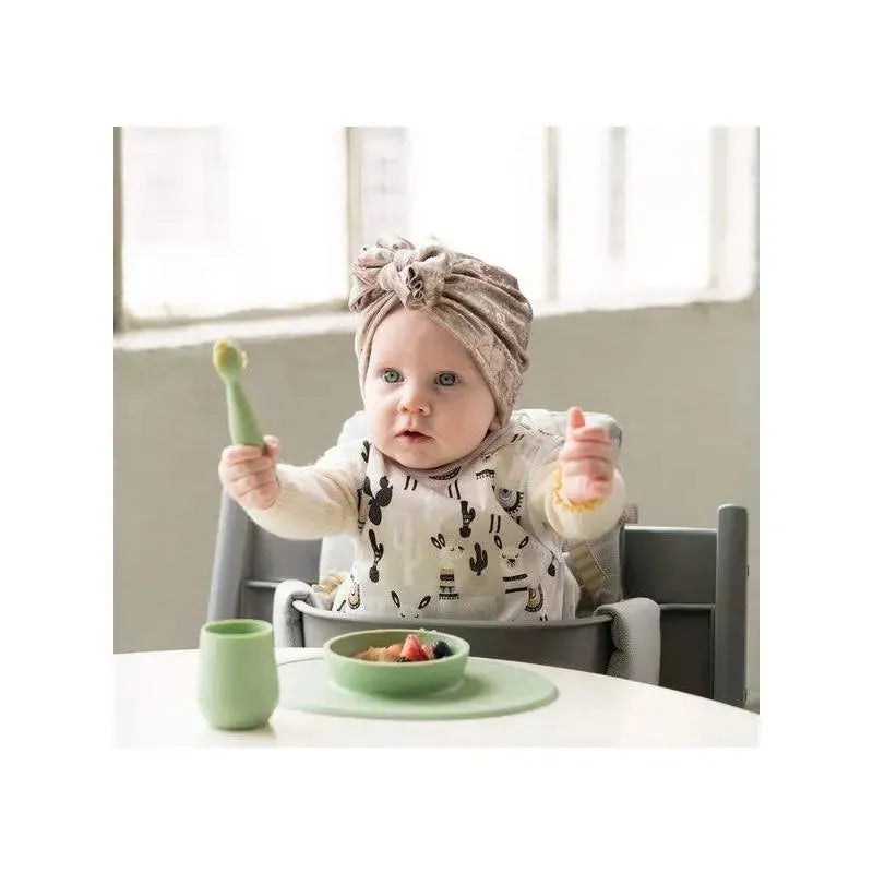 A baby in a high chair with a sage Ezpz Tiny Spoon holds a spoon up while food is on a green plate in front.