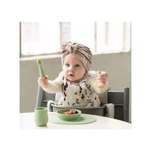 A baby in a high chair with a sage Ezpz Tiny Spoon holds a spoon up while food is on a green plate in front.