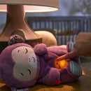 Purple monkey plush sound machine with a glowing belly, being activated by a hand, with a soft lit ambiance.