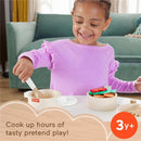 Child in a lavender shirt joyfully pretends to cook with a Fisher Price wooden pots and pans set, mixing colorful food items.
