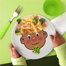 Ceramic dinner plate featuring a cartoon face design, topped with pasta, broccoli, and hot dog slices, held by a child.