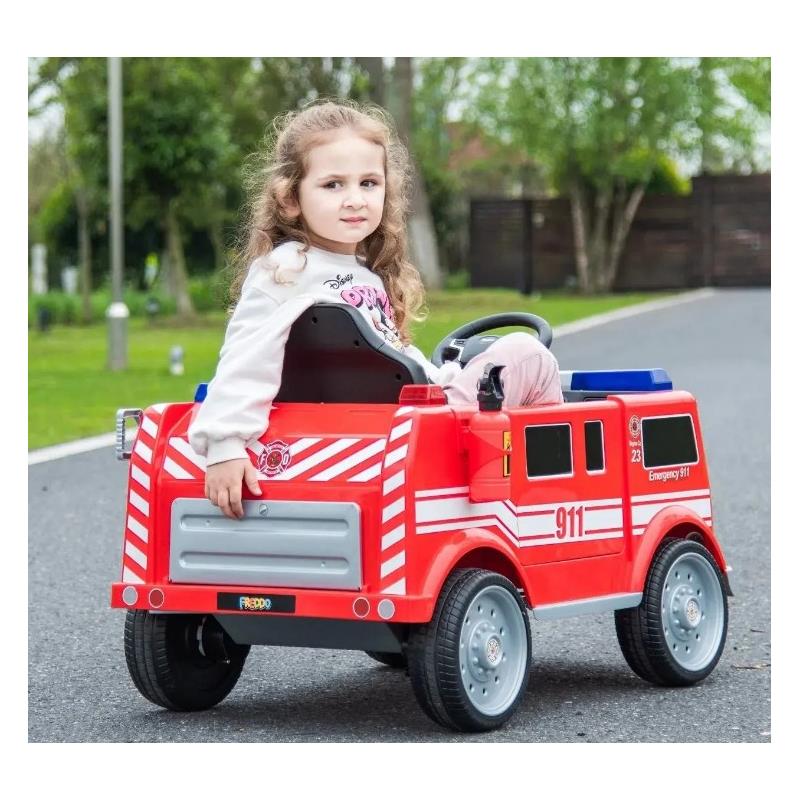 Child sitting in a red Freddo fire truck toy, with emergency markings, on a driveway surrounded by greenery.