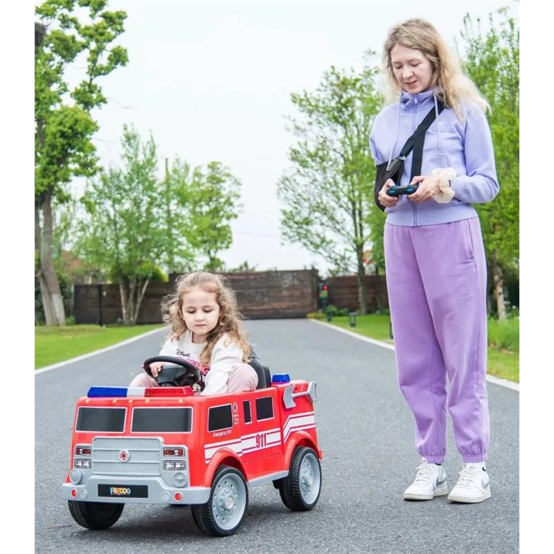A child sits in a red, plastic Freddo fire truck toy on a paved path, while an adult supervises nearby.