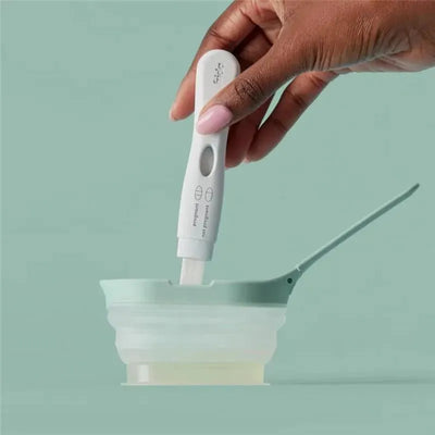 A hand holds a Frida Fertility Early Detection Pregnancy Test over a small collection cup, designed for urine samples.