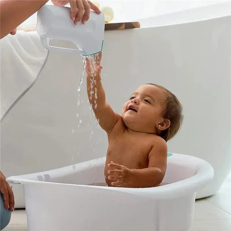 A baby in a tub enjoys water being poured from a Fridababy Control The Flow Rinser by an adult.