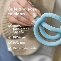 Muted blue FridaBaby Get-A-Grip teether held in a hand, featuring text on safety and cleaning features in the background.