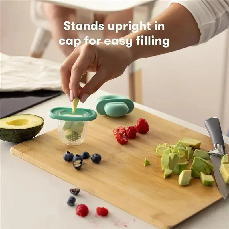 Teal Fridababy Push Pop Feeder held by a hand, positioned on a cutting board with cut fruit, featuring a pile of chopped avocado, raspberries, and blueberries.