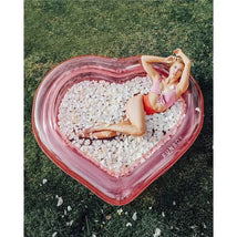 Giant inflatable clear pink heart kiddie pool by Funboy, filled with white petals, a woman lounging in swimwear.