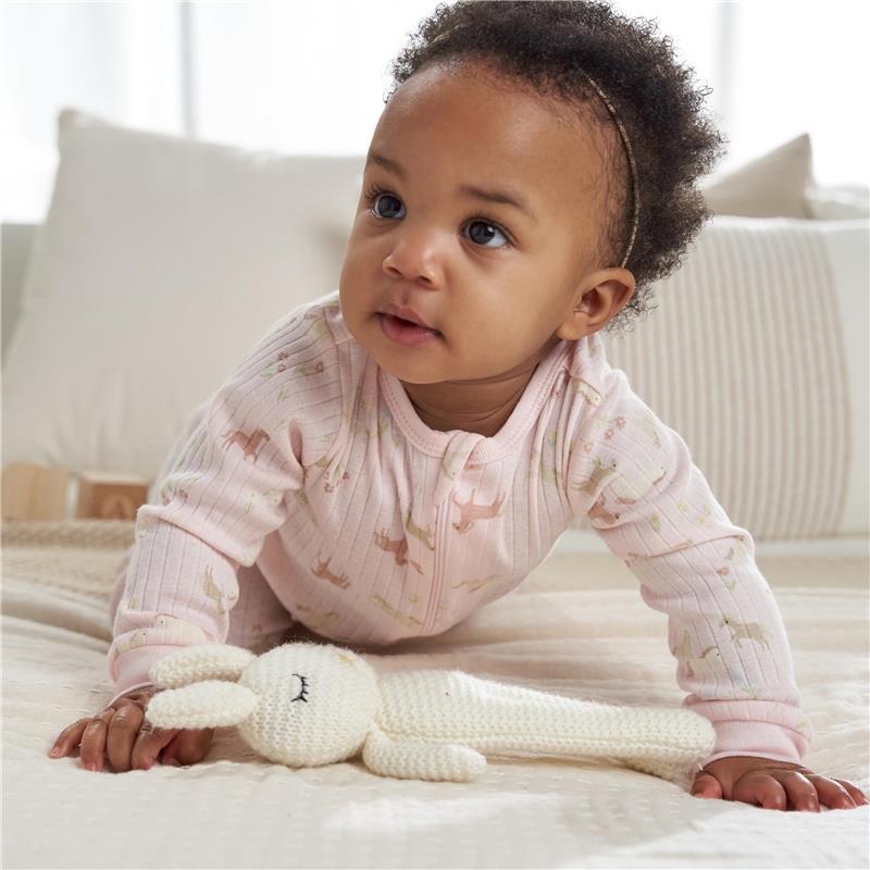 Baby crawling on a soft bed, wearing a pink ribbed sleep suit with playful animal prints, next to a knitted bunny toy.