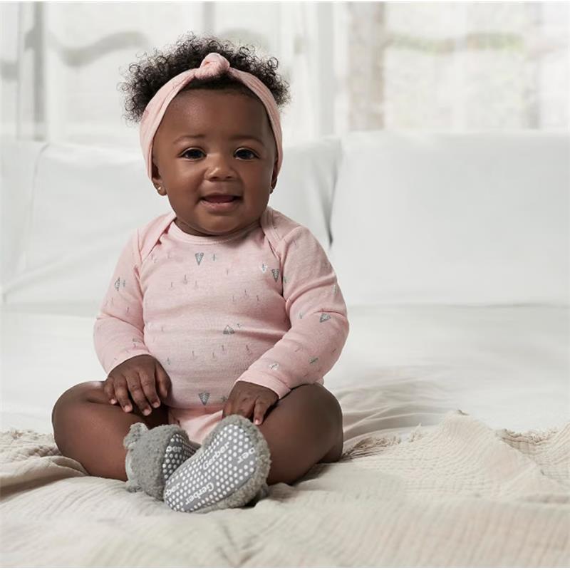 Baby girl sitting on a bed wearing a pink long sleeve onesie with silver patterns, gray booties, and a matching headband.