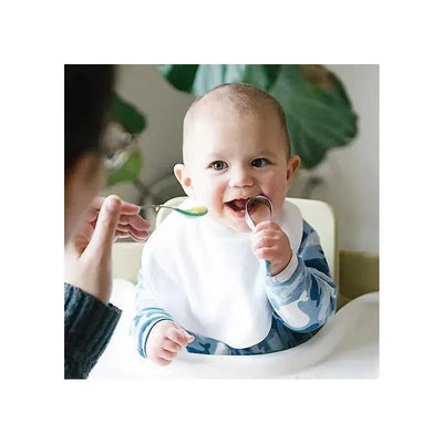 Baby wearing a white bib, sitting in a high chair, grinning while holding two spoons during mealtime.