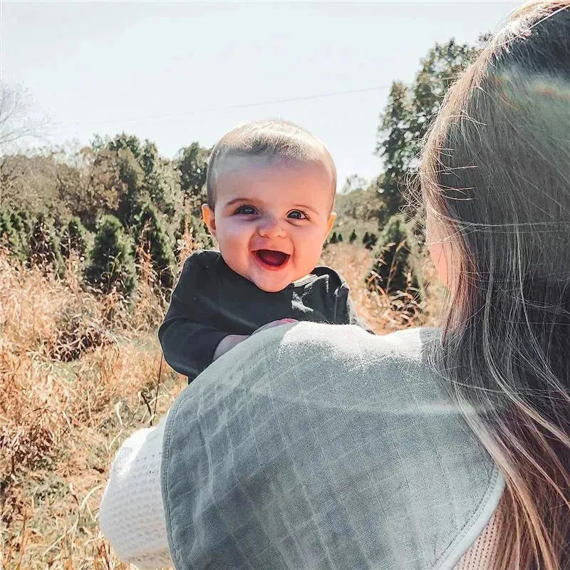Smiling baby held by an adult, outdoors in a field, wearing a black shirt and surrounded by nature.
