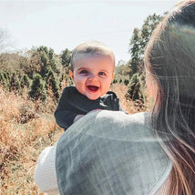 Smiling baby held by an adult, outdoors in a field, wearing a black shirt and surrounded by nature.