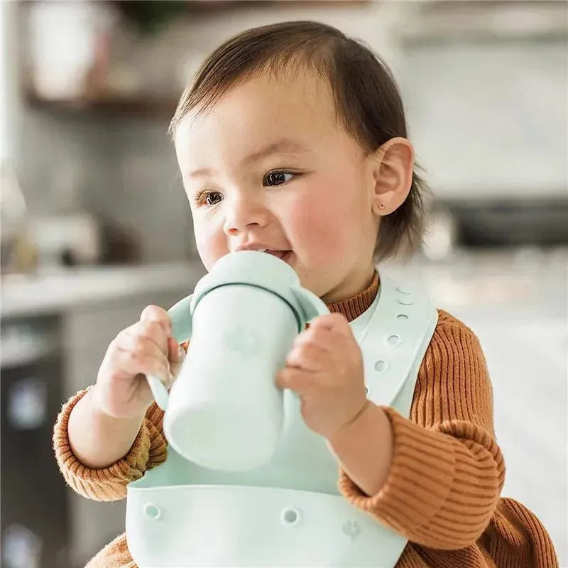 Toddler wearing a bib holds a mint green sip and straw cup while smiling, set in a kitchen environment.