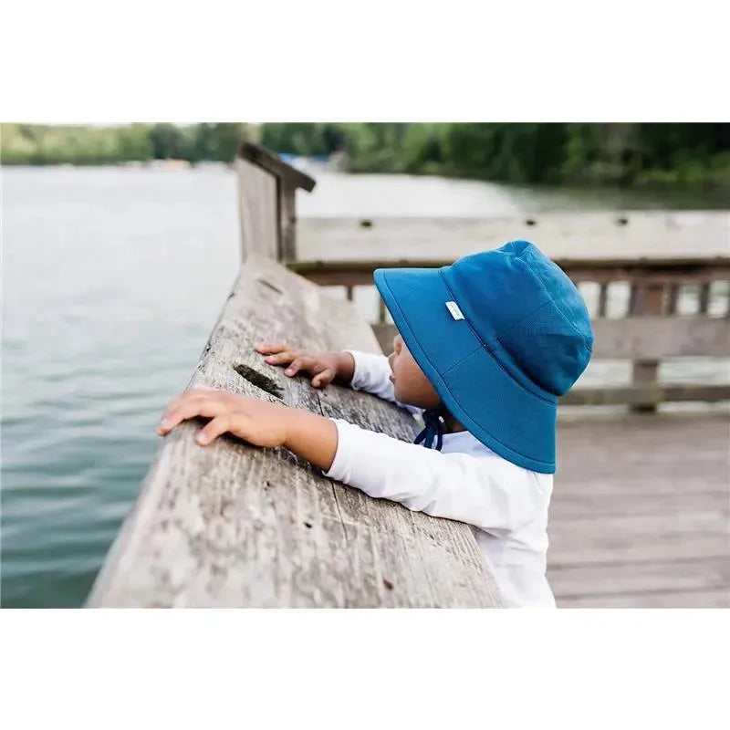 Baby wearing a navy Green Sprouts breathable swim and sun bucket hat, leaning over a wooden railing by a lake.