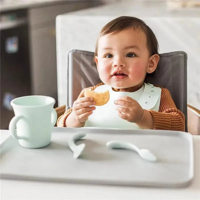 Toddler sitting in a high chair, holding a snack, with a Green Sprouts light grapefruit silicone learning cup nearby.