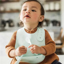 Toddler wearing a light blue bib and holding a spoon, looking attentively, with a soft background hinting at a kitchen setting.