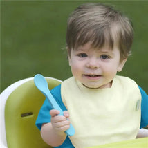 Infant boy wearing a yellow bib, holding a blue spoon, seated in a high chair, smiling outdoors.