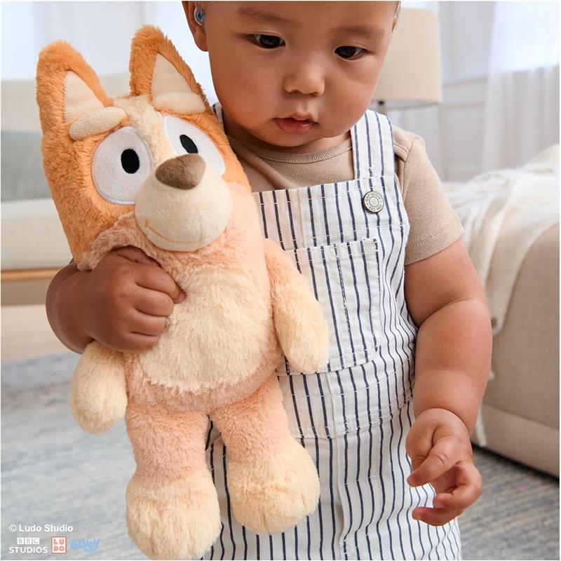 Child holding a soft, orange Bingo plush toy, wearing striped overalls, with a neutral background.