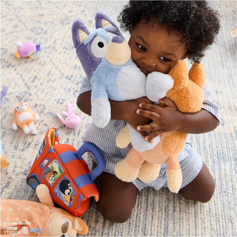 Child hugging two plush toys, one blue and one orange, surrounded by additional plush toys and a colorful backpack.
