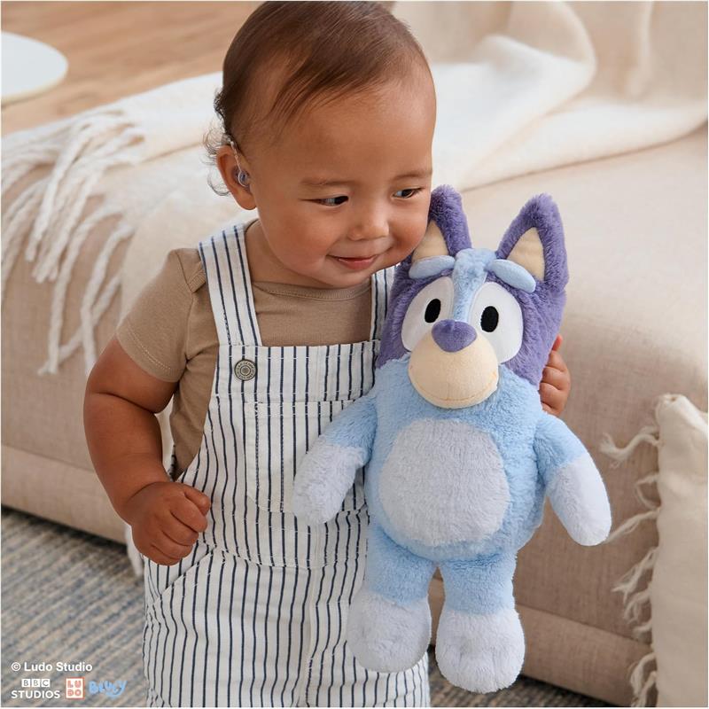 A toddler in striped overalls smiles while holding a soft blue plush toy resembling Bluey, a cartoon character.