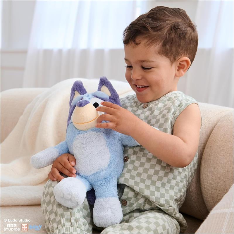 Child playing with a blue plush toy resembling Bluey, smiling while seated on a couch in a bright room.