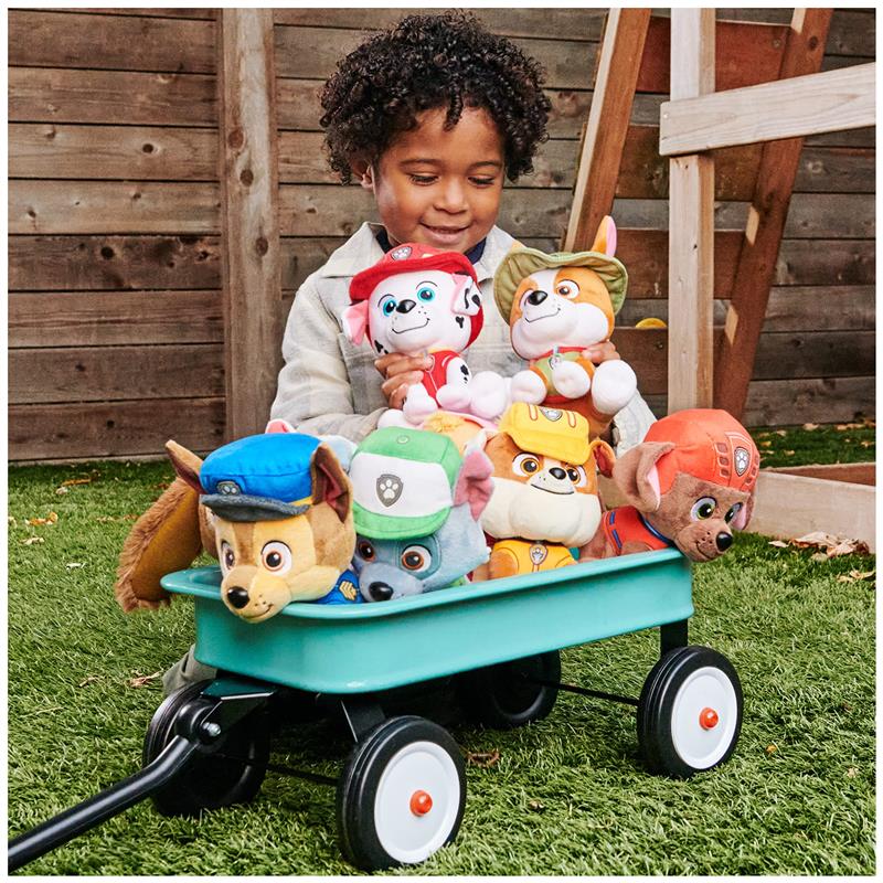 A child smiles while holding plush PAW Patrol characters in a green wagon on grass.