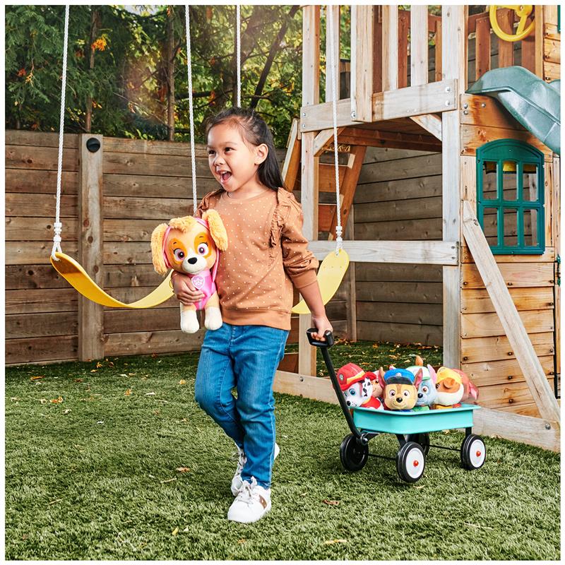 Girl smiling and holding a Plush PAW Patrol Zuma toy, pulling a wagon with other plush characters in a playground.