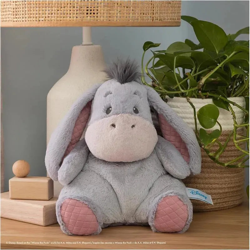 Soft gray plush toy of Eeyore, featuring large ears and a friendly expression, placed beside a lamp and a potted plant.