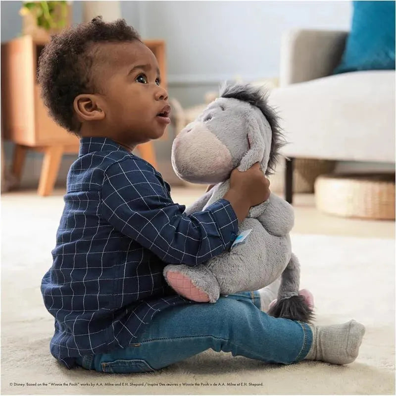 Toddler in a blue checkered shirt cuddles a soft plush Eeyore toy while sitting on a light-colored rug.