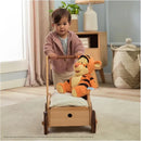 Toddler pushing a wooden toy cart with a plush orange Tigger toy sitting on a white blanket.
