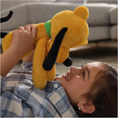 A child smiles while lying on the floor, joyfully holding a plush yellow dog toy with black ears, named Pluto.